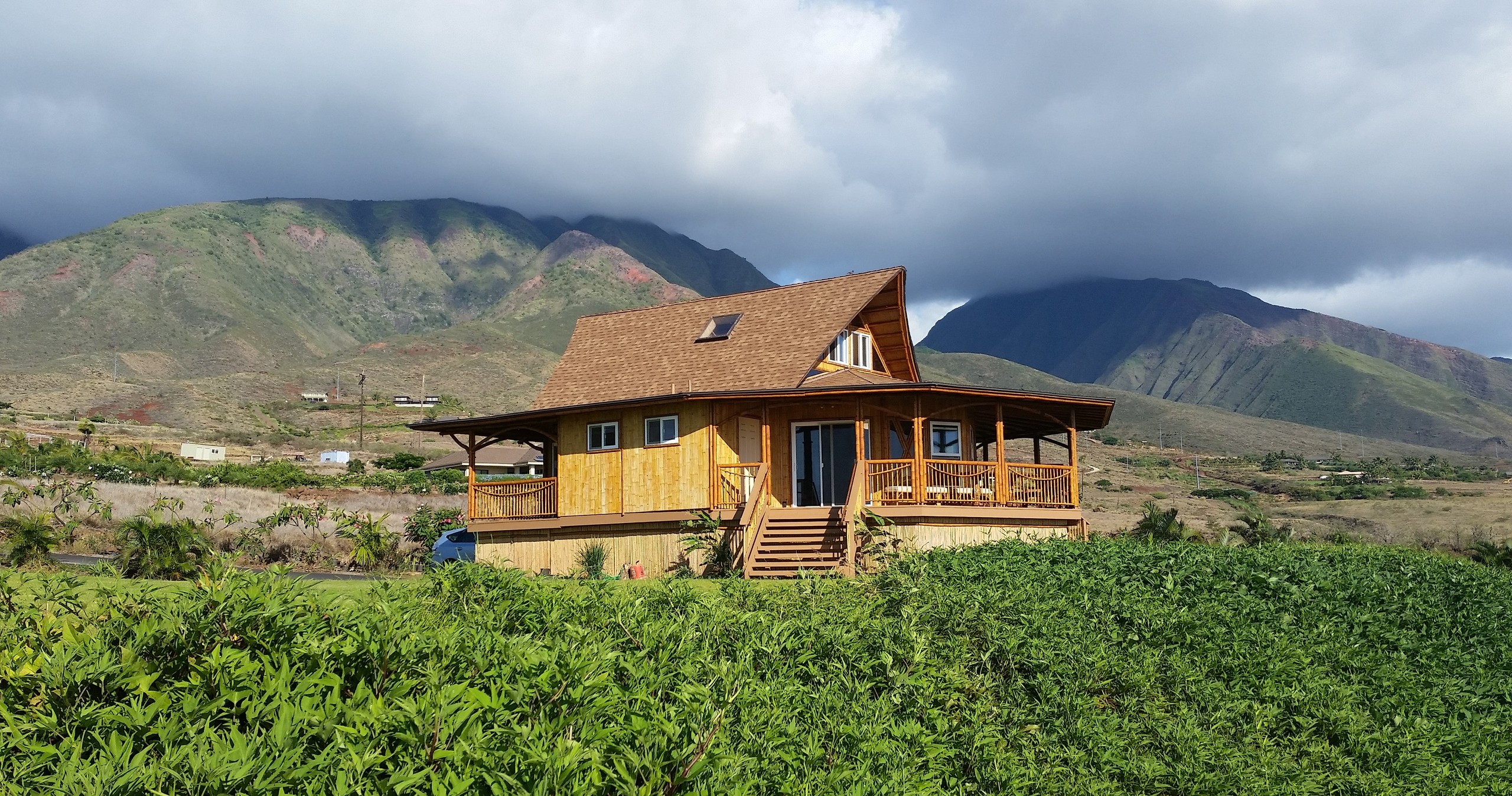 Wooden house with porch set against mountains under cloudy sky, surrounded by green vegetation.
